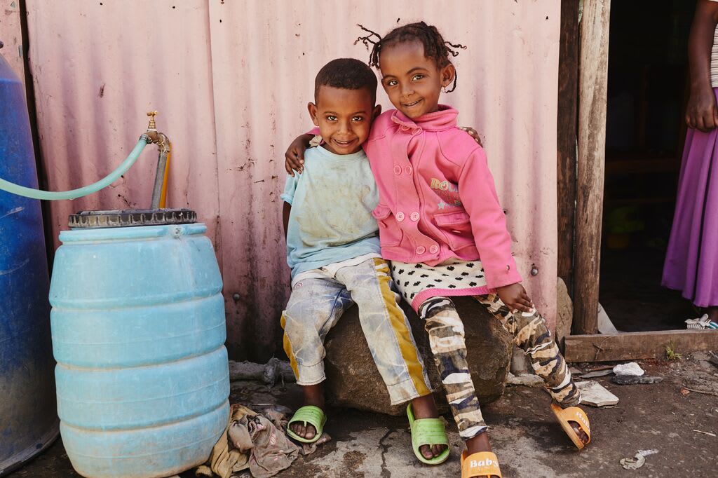 One little girl and one little boy smile with their arms around eachother beside a rainwater filter.